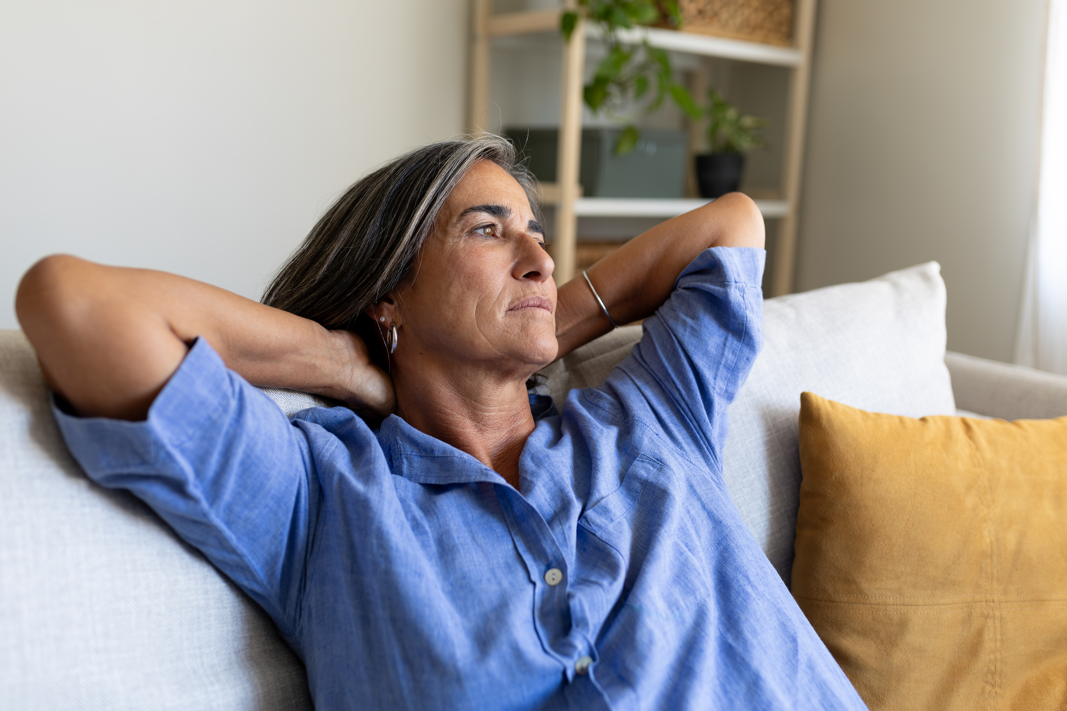 Serene or thoughtful mature woman rests on a comfortable sofa, hands behind her head.