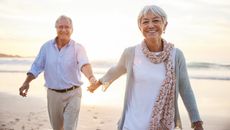 Portrait of a laughing senior woman leading her husband by the hand while walking on a sandy beach at sunset