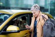A senior woman with a duffel bag checks her phone before getting into a car.