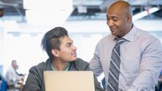 A financial professional smiles as he mentors a young man who's using a laptop.