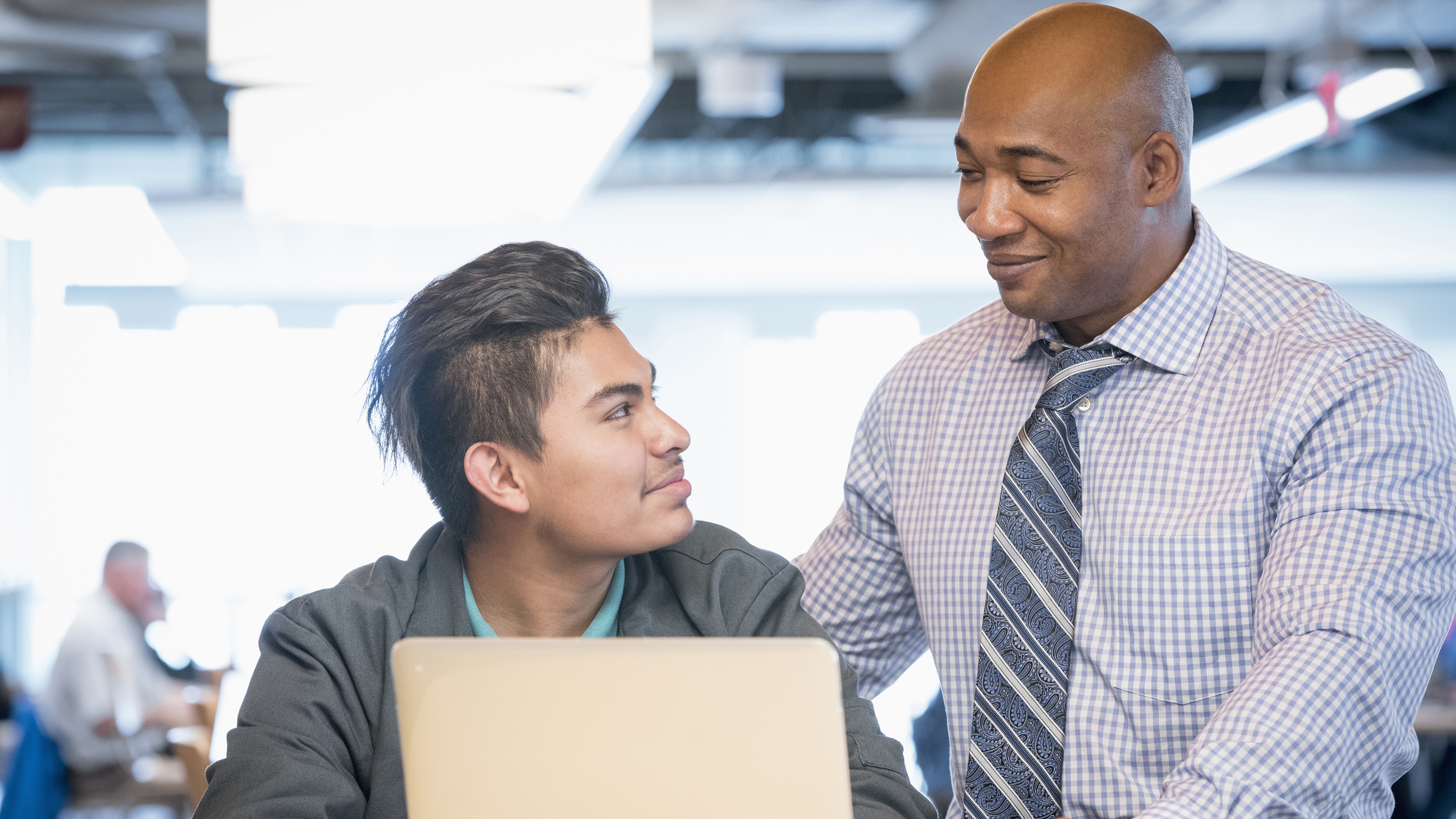 A financial professional smiles as he mentors a young man who's using a laptop.