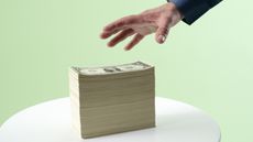 A man's hand reaches for a very tall stack of cash.