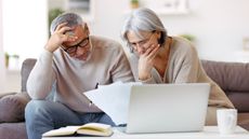 A confused-looking retired couple look over paperwork on their living room sofa.